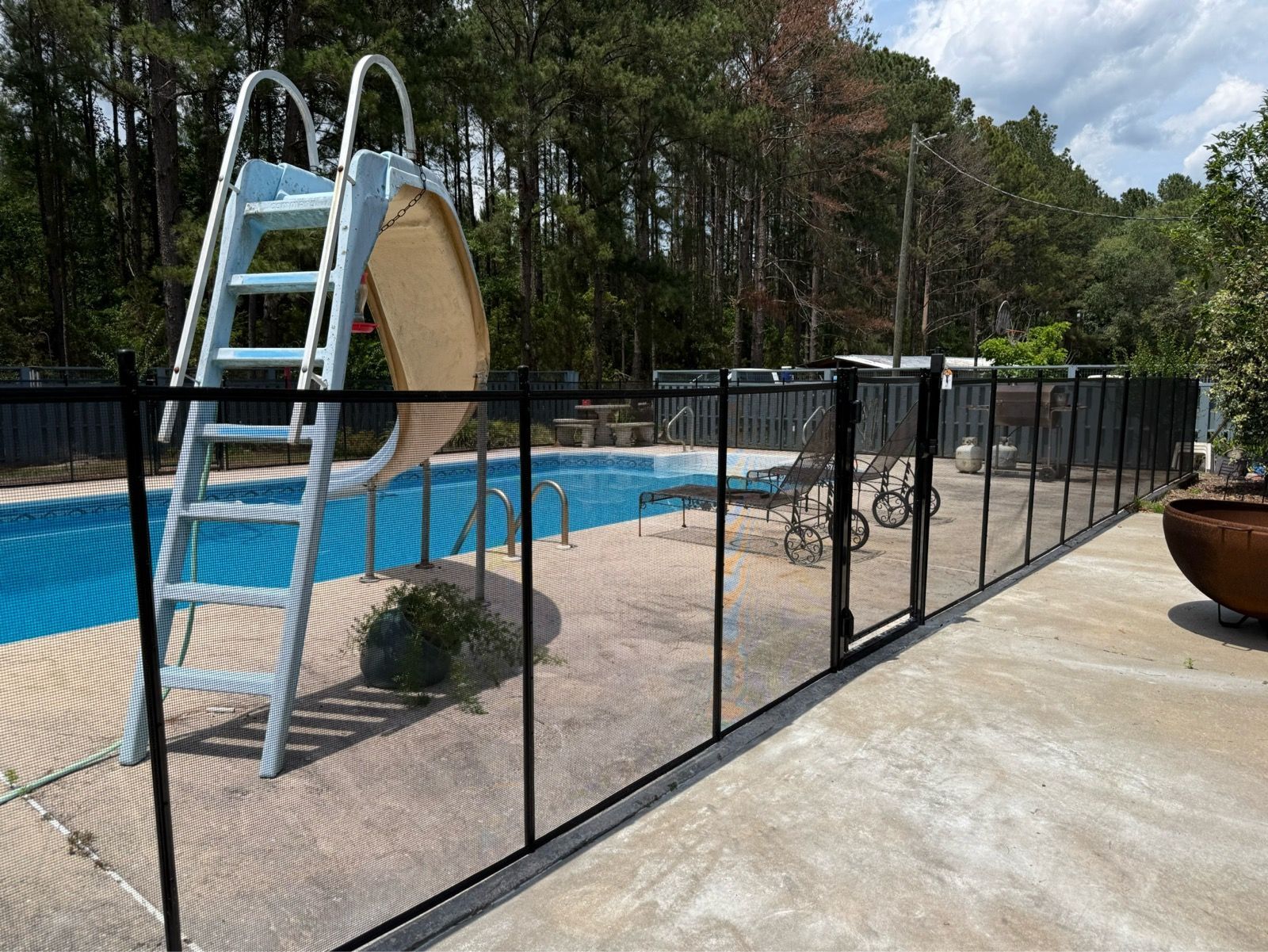 Pool with metal slide and black fence beside a concrete patio and trees.