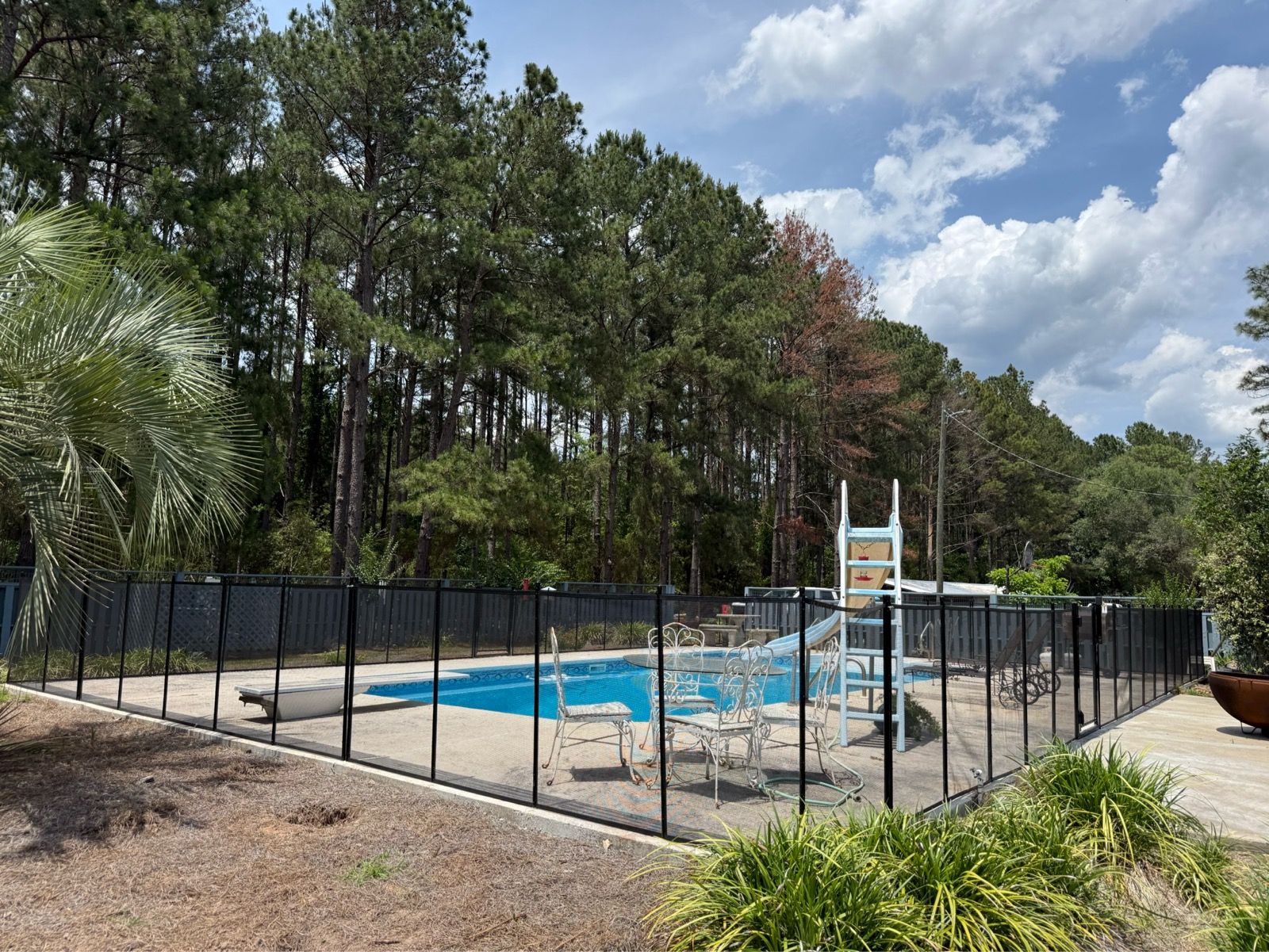 Fenced outdoor swimming pool with blue water, white diving board, and trees under a partly cloudy sky.