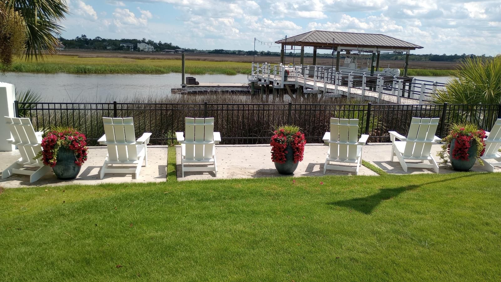 White chairs on a lawn facing a waterfront gazebo, with flower arrangements along the aisle.