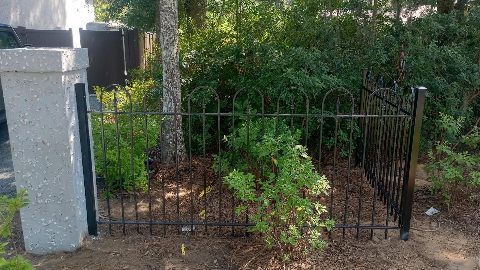 Black metal gate and aluminum fence beside a stone post, with green shrubs and trees behind it.