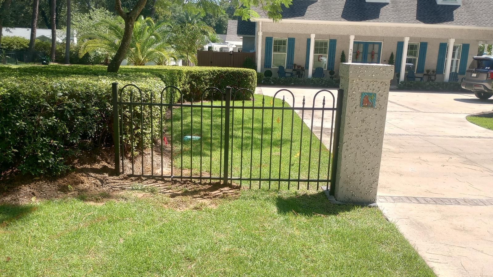 Front yard with a black fence, stone post, green grass, and a house in the background.