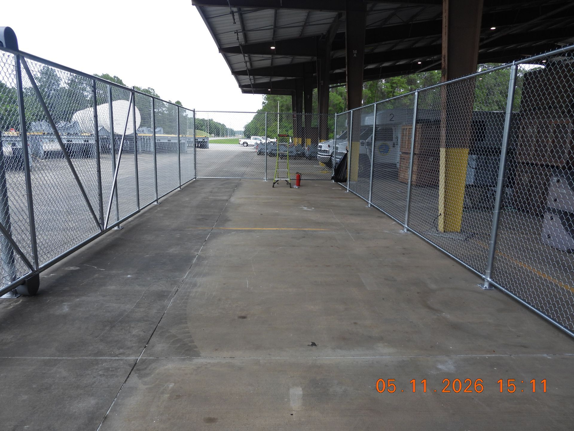 Covered sidewalk beside a chain-link fence and parking lot, with a few orange cones ahead.