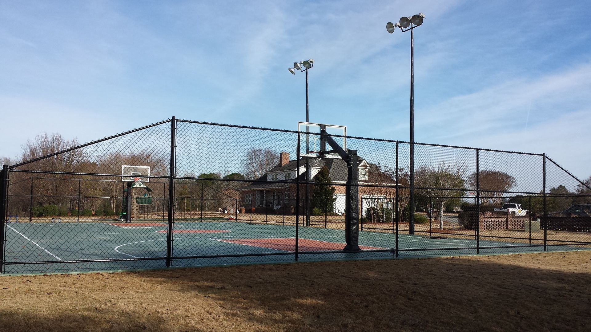 Outdoor tennis court behind a chain-link fence, with lights, net, and bare trees under a clear sky.