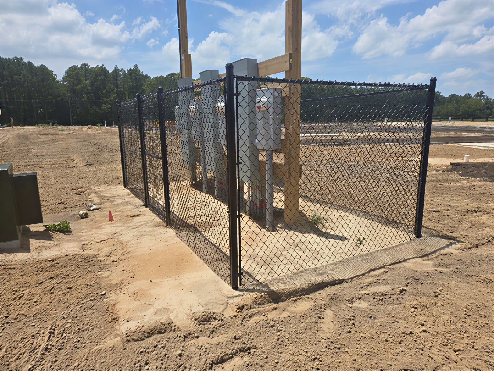 Chain-link fenced utility enclosure on sandy construction site with a tall pole and control box.