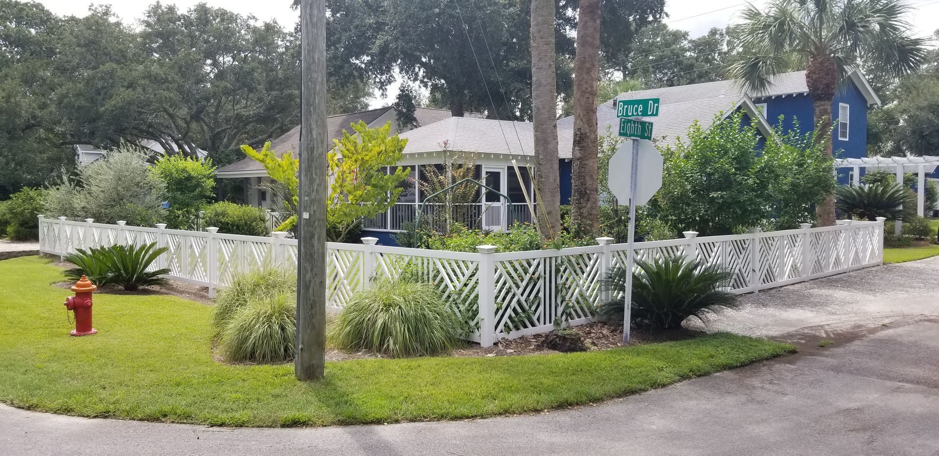 A white lattice fence surrounds a house and garden on a corner lot with a fire hydrant and a street sign nearby.