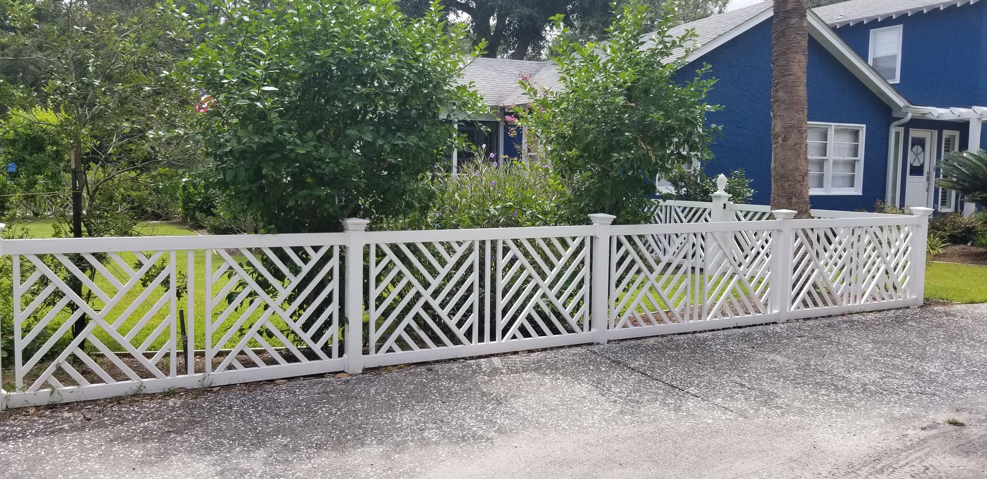 White fence with an intricate, geometric diagonal lattice pattern in front of a blue house and green trees.