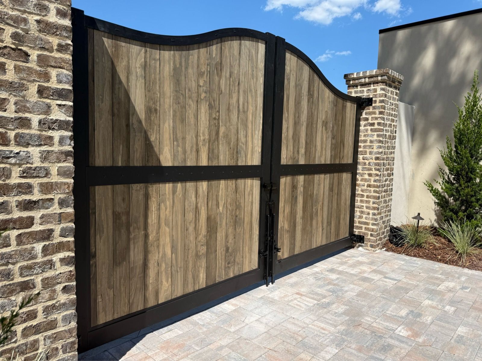 Wooden driveway gate with black frame beside brick pillars and a paved courtyard under a blue sky