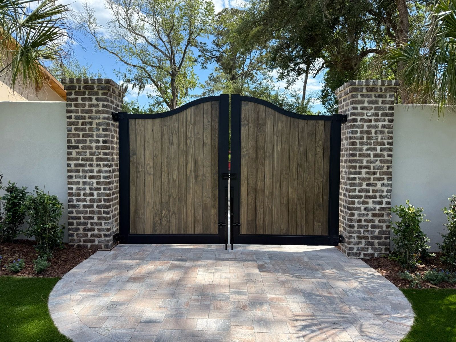 Wooden driveway gate with brick pillars and a curved concrete driveway, framed by trees and landscaping