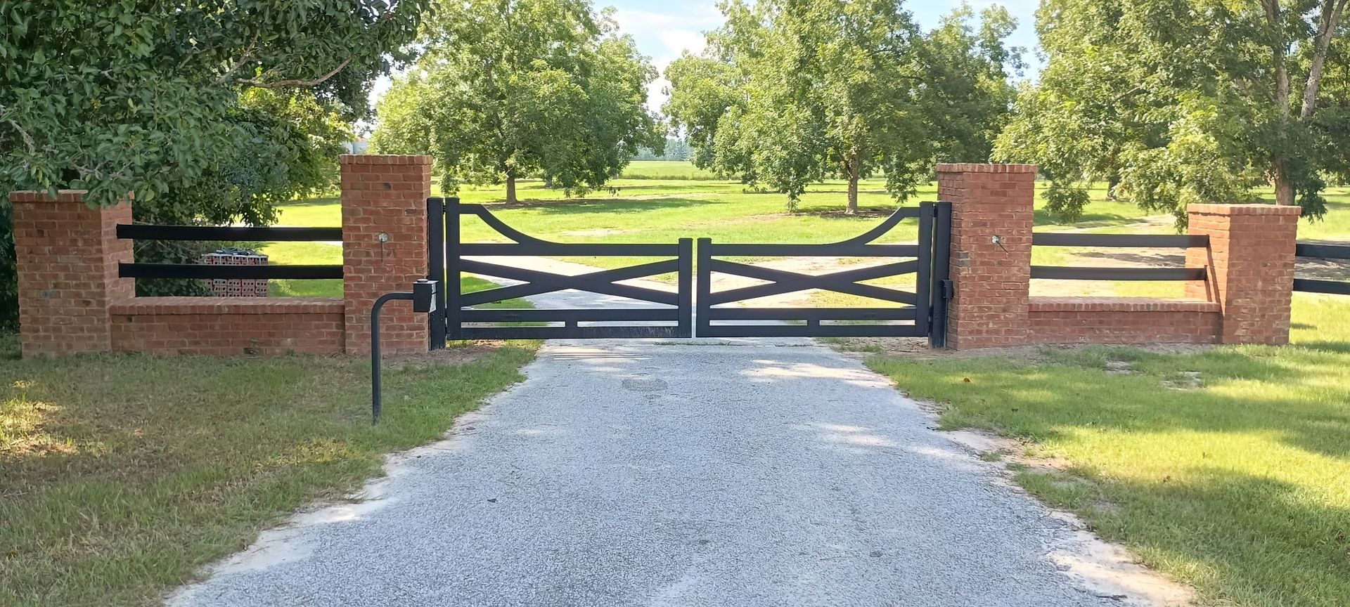 A brick pillar and black gate entrance to a grassy drive with trees.