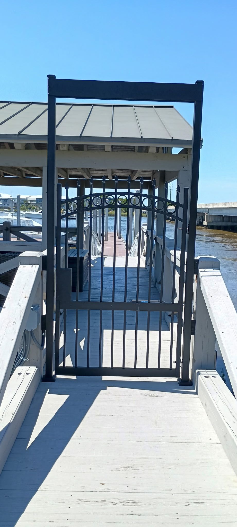 A metal gate on a wooden dock leads to a covered walkway over water under a blue sky.