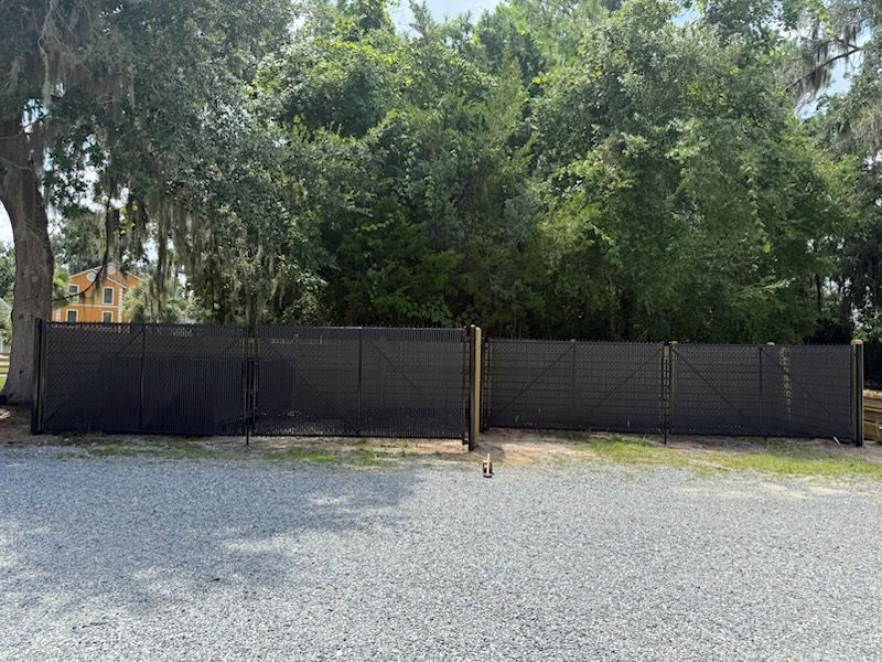 Black wooden fence in front of trees and gravel ground. A gate is in the middle.