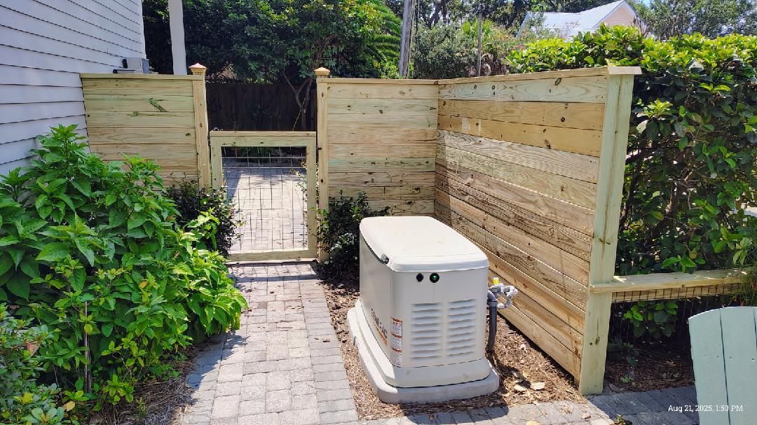 Generator enclosed in a wooden fence with a gated walkway, located in a yard.