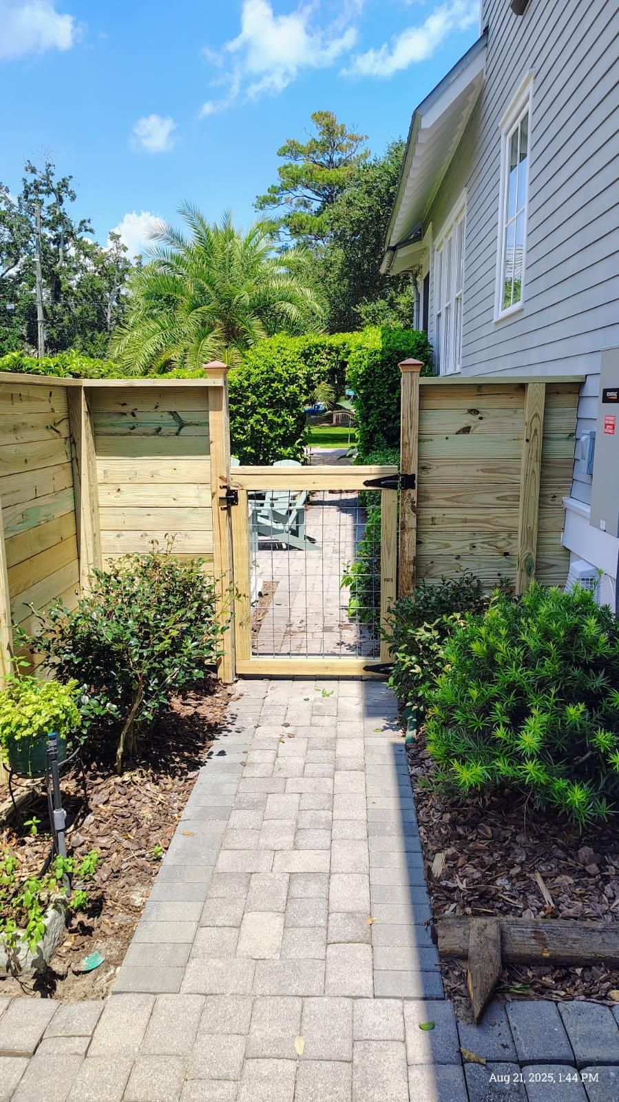 Wooden fence with gate and paved path leading to it. Green bushes and side of a light gray house.