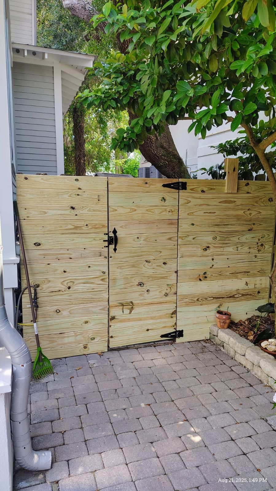 Wooden gate in a brick-paved yard, between a white building and a tree.