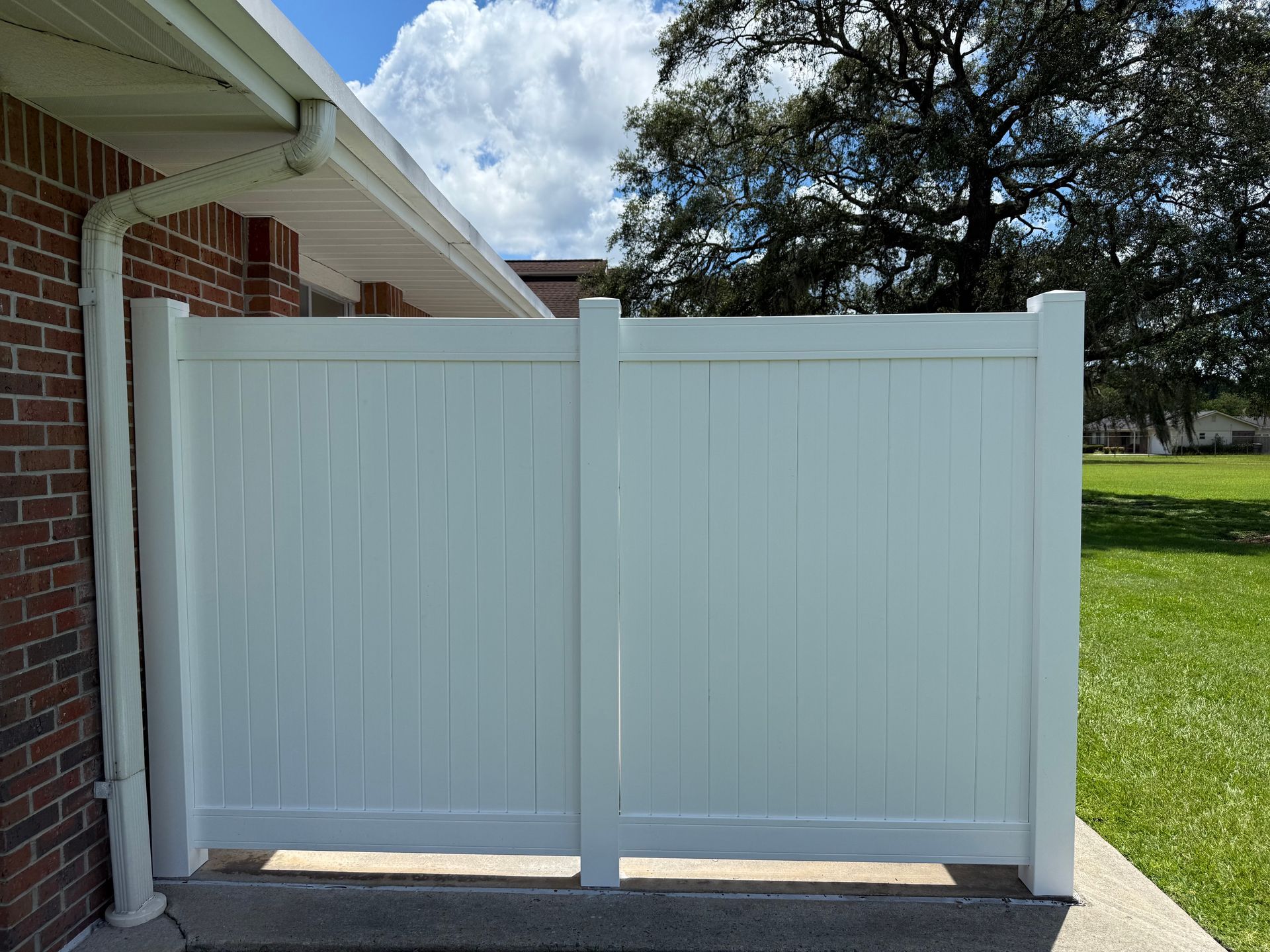 White vinyl fence next to a brick building with green grass and a blue sky background.