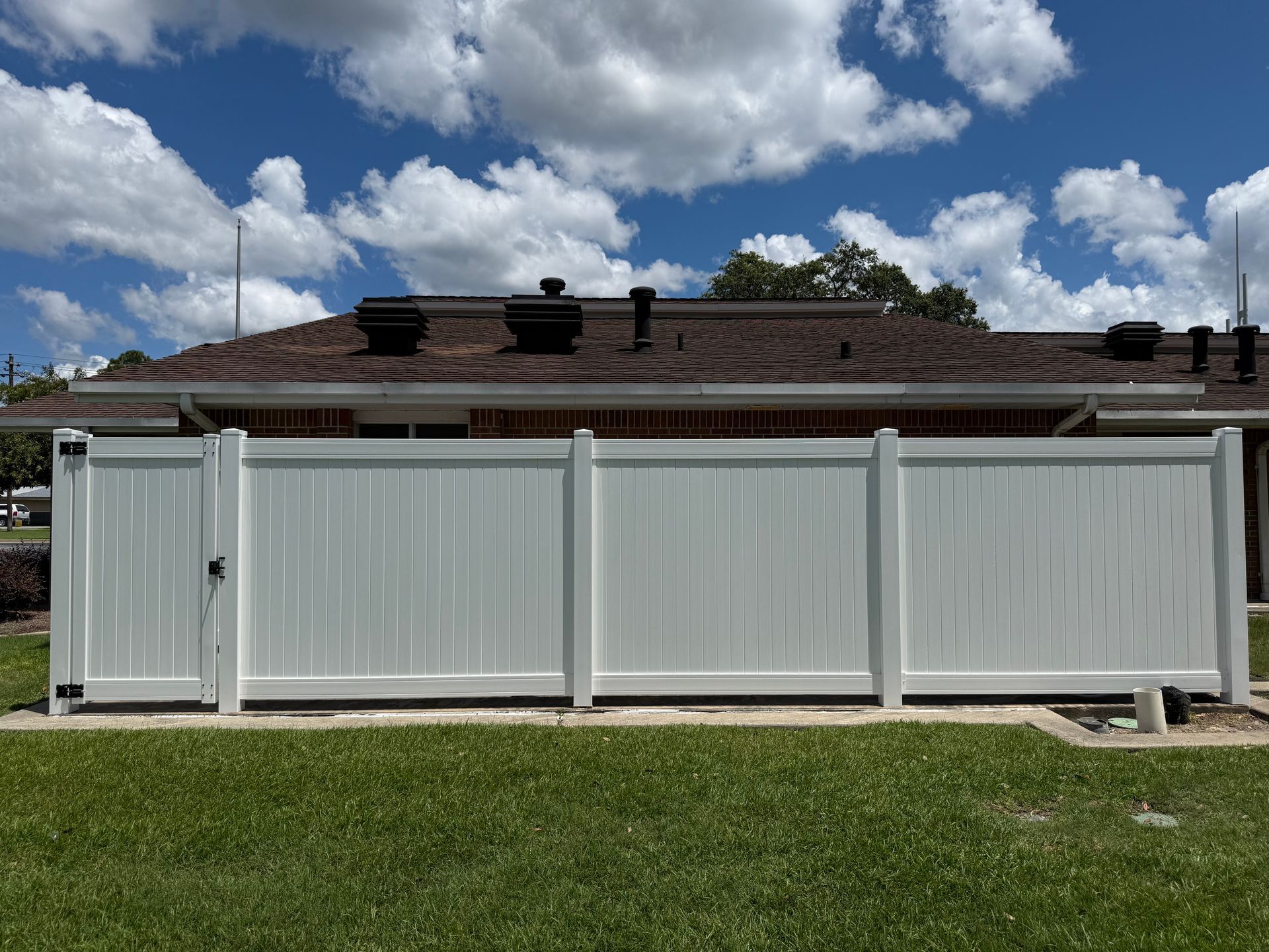 White vinyl fence and gate in a grassy yard, behind a brown roof against a partly cloudy blue sky.