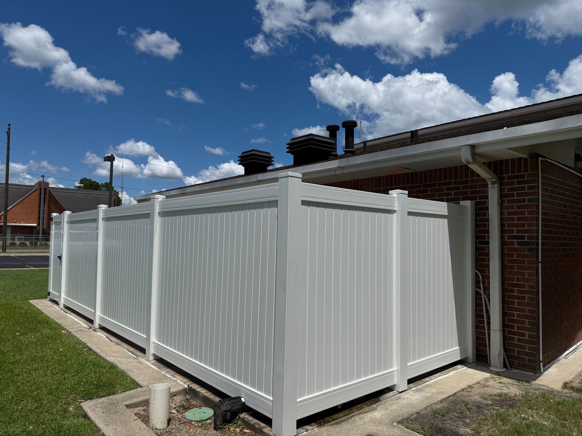 White vinyl fence next to a brick building, under a blue sky with clouds.