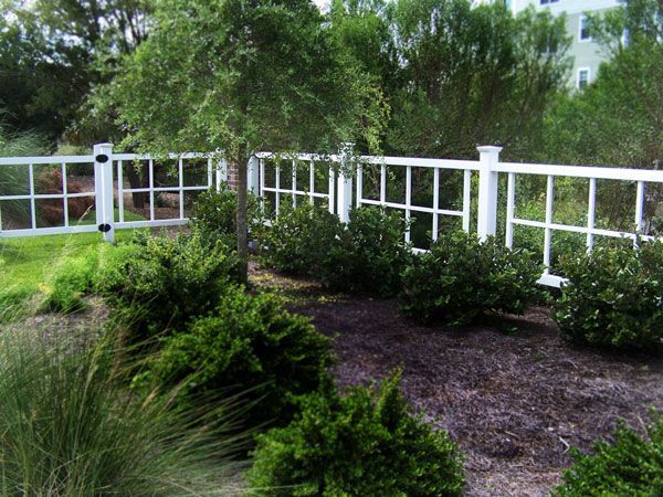White grid-style fence in front of green bushes and trees. The fence borders a grassy area.