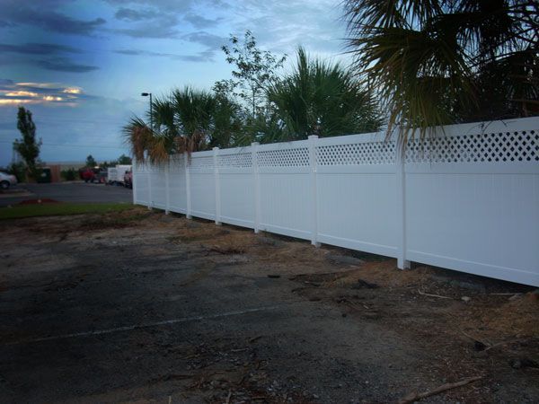 White vinyl fence bordering a parking lot, under a cloudy sky. The top portion of the fence has a lattice design.