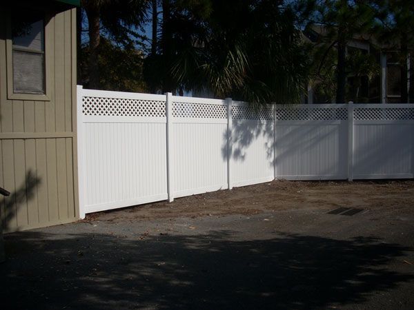 White vinyl fence with lattice top section, bordering a property with trees and building.