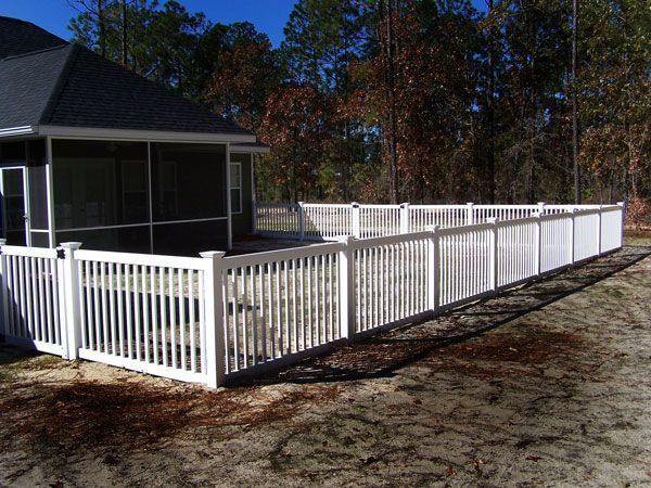 White picket fence surrounding a house with a screened porch, set in a wooded area under a blue sky.