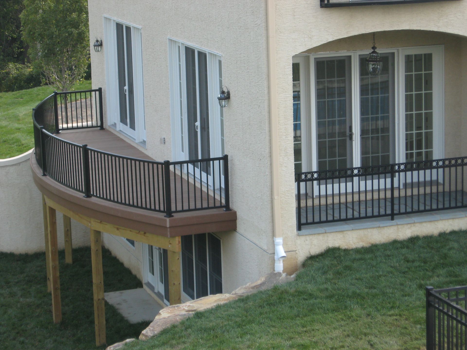 Curved deck with black railing attached to a tan stucco house with white doors and a grassy yard.