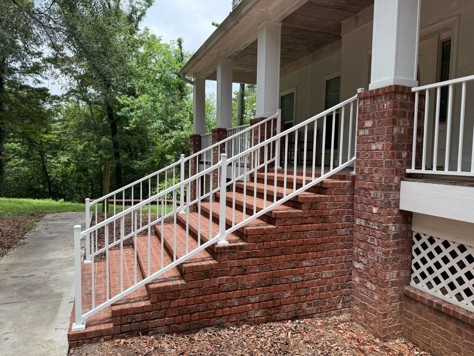 Brick house porch with white railing and steps leading up from a concrete walkway.