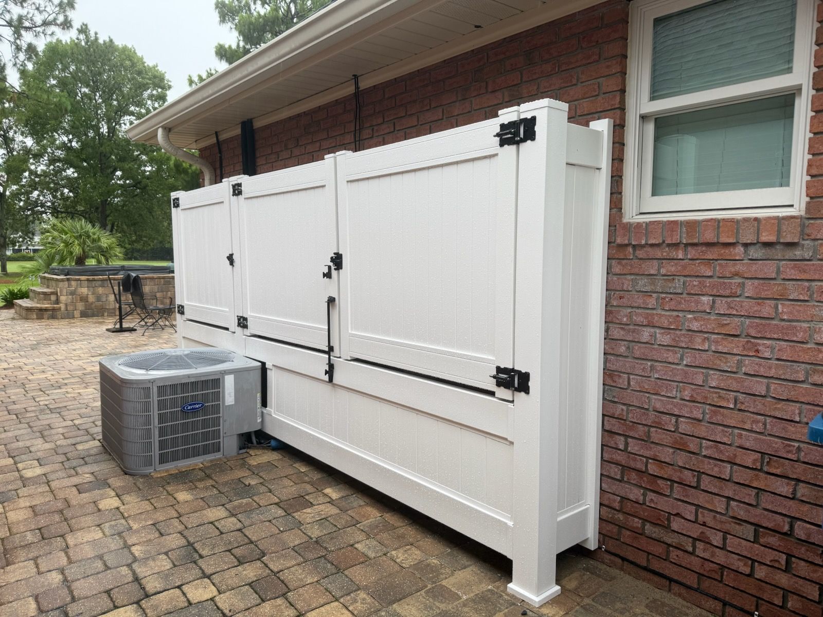A white vinyl fence with access doors encloses a central air conditioning unit beside a brick building.