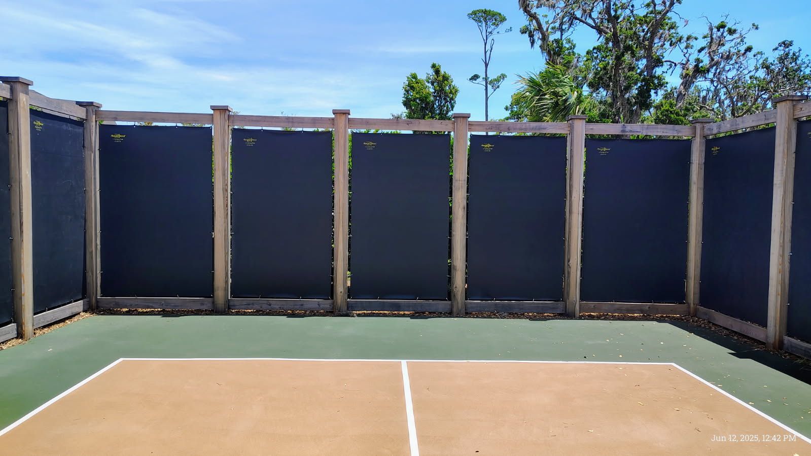 Pickleball court surrounded by black privacy screens and wooden posts, against a blue sky.