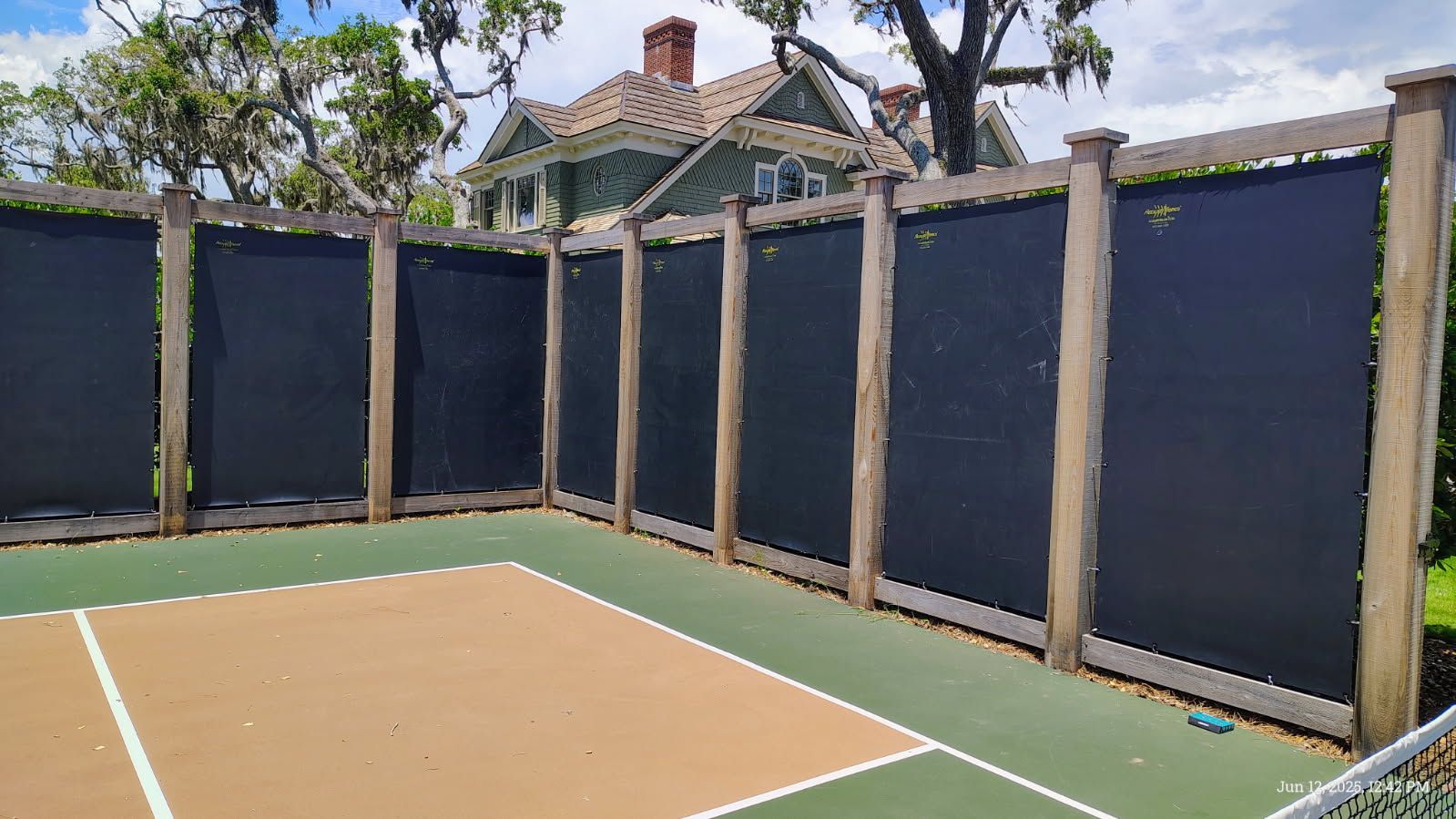 A pickleball court with a dark privacy fence, a two-toned surface, and a green building in the background.