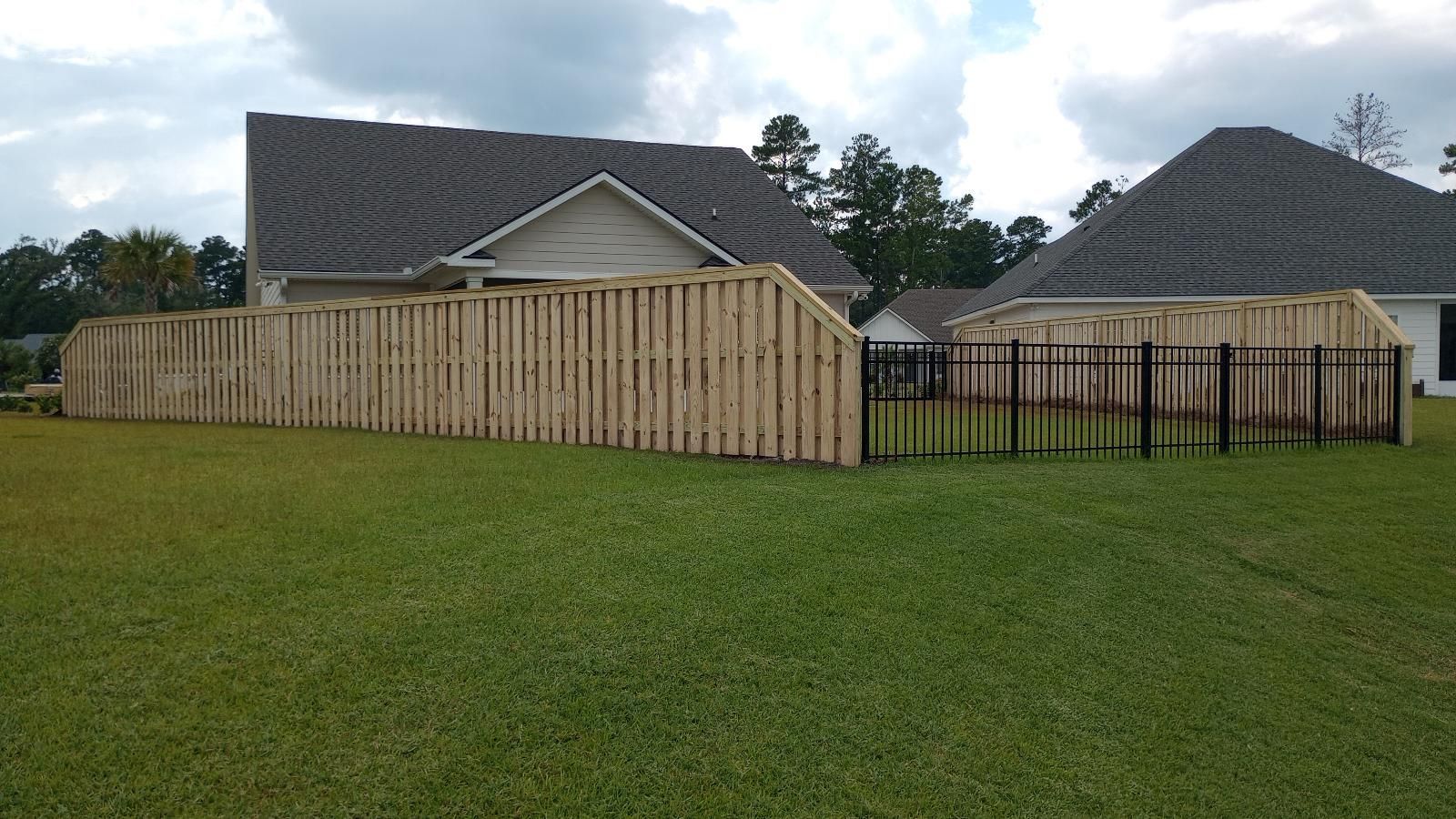 Wooden privacy fence around a suburban backyard with green lawn and two beige houses under cloudy sky