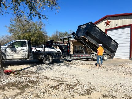 Truck unloading dumpster trailer on a gravel driveway beside a red-roofed building. A person watches.