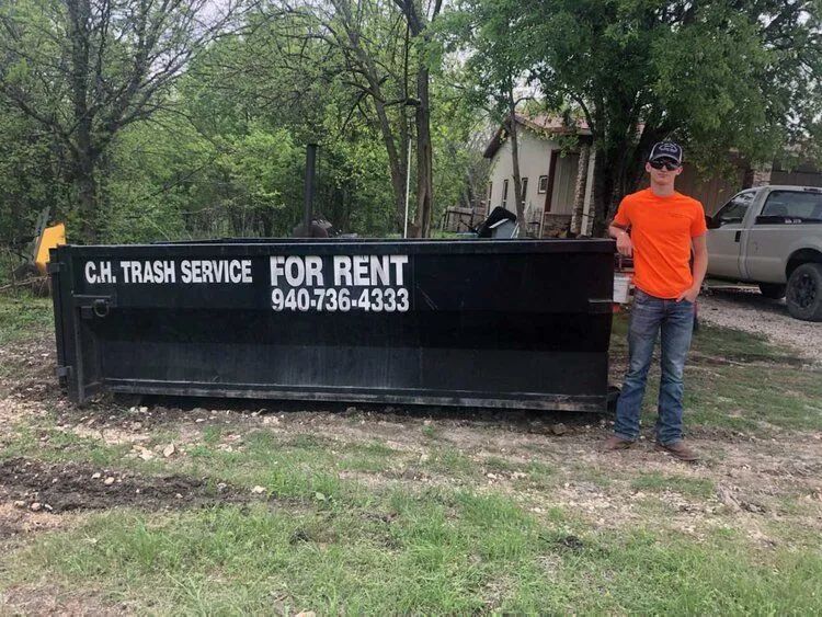Man in orange shirt stands beside a black dumpster labeled 
