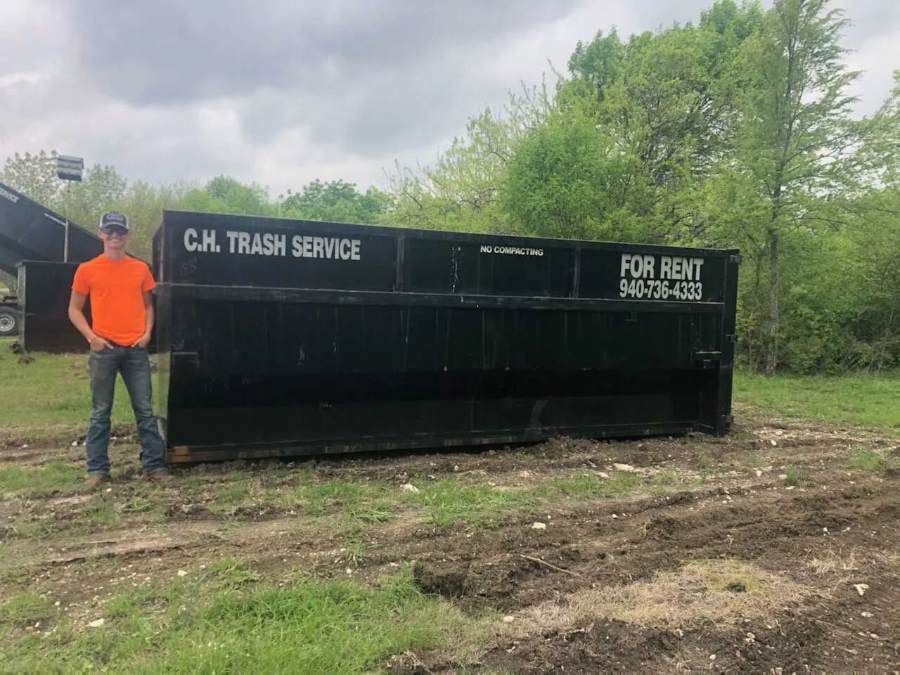 A person stands beside a large, black dumpster with 