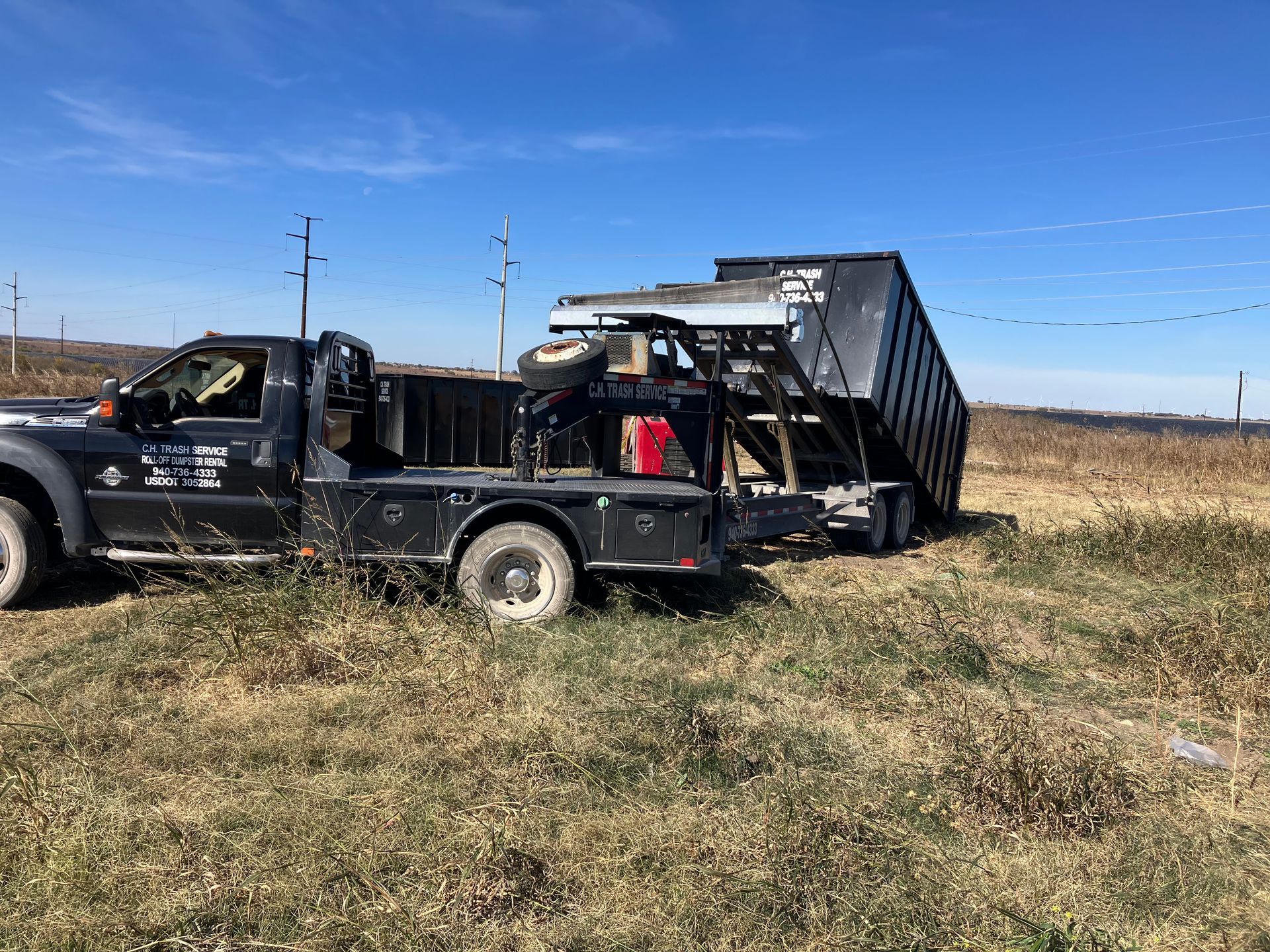 Black truck with a trailer hauling a black dumpster on a grassy field under a blue sky.