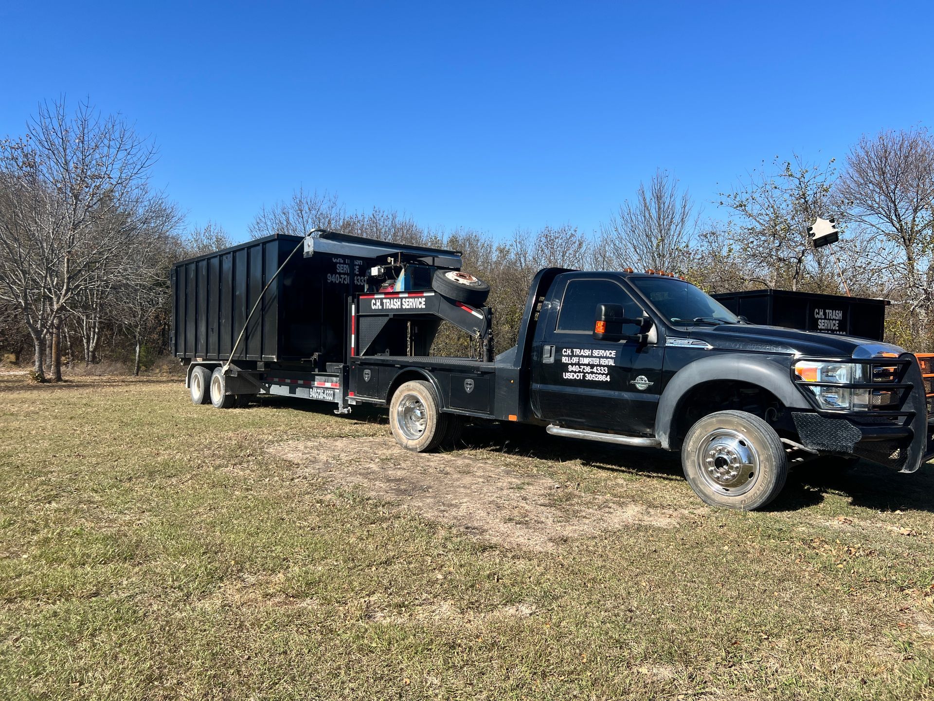 Black truck towing a black dumpster trailer on a grassy field under a blue sky.