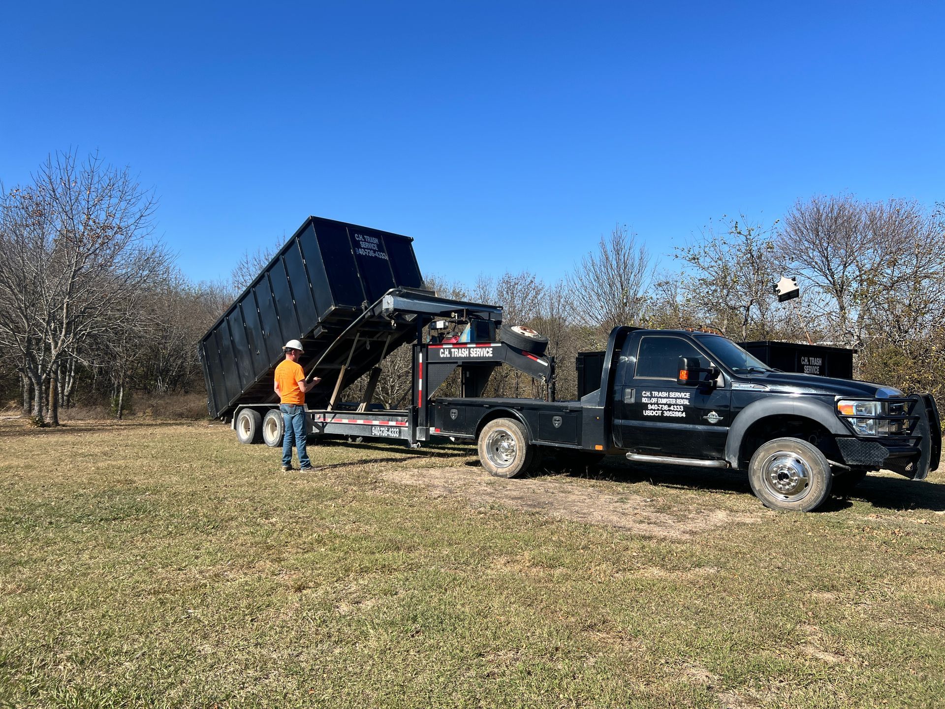 Truck with a raised dumpster trailer on a grassy field, under a clear blue sky. A person stands nearby.