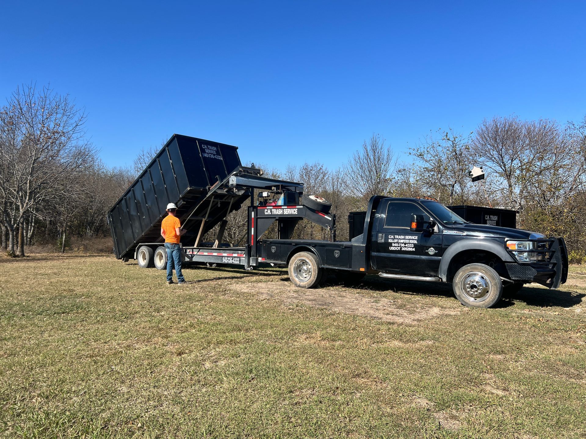 Truck unloading a large, black dumpster onto a grassy field on a sunny day. A person in an orange vest stands nearby.