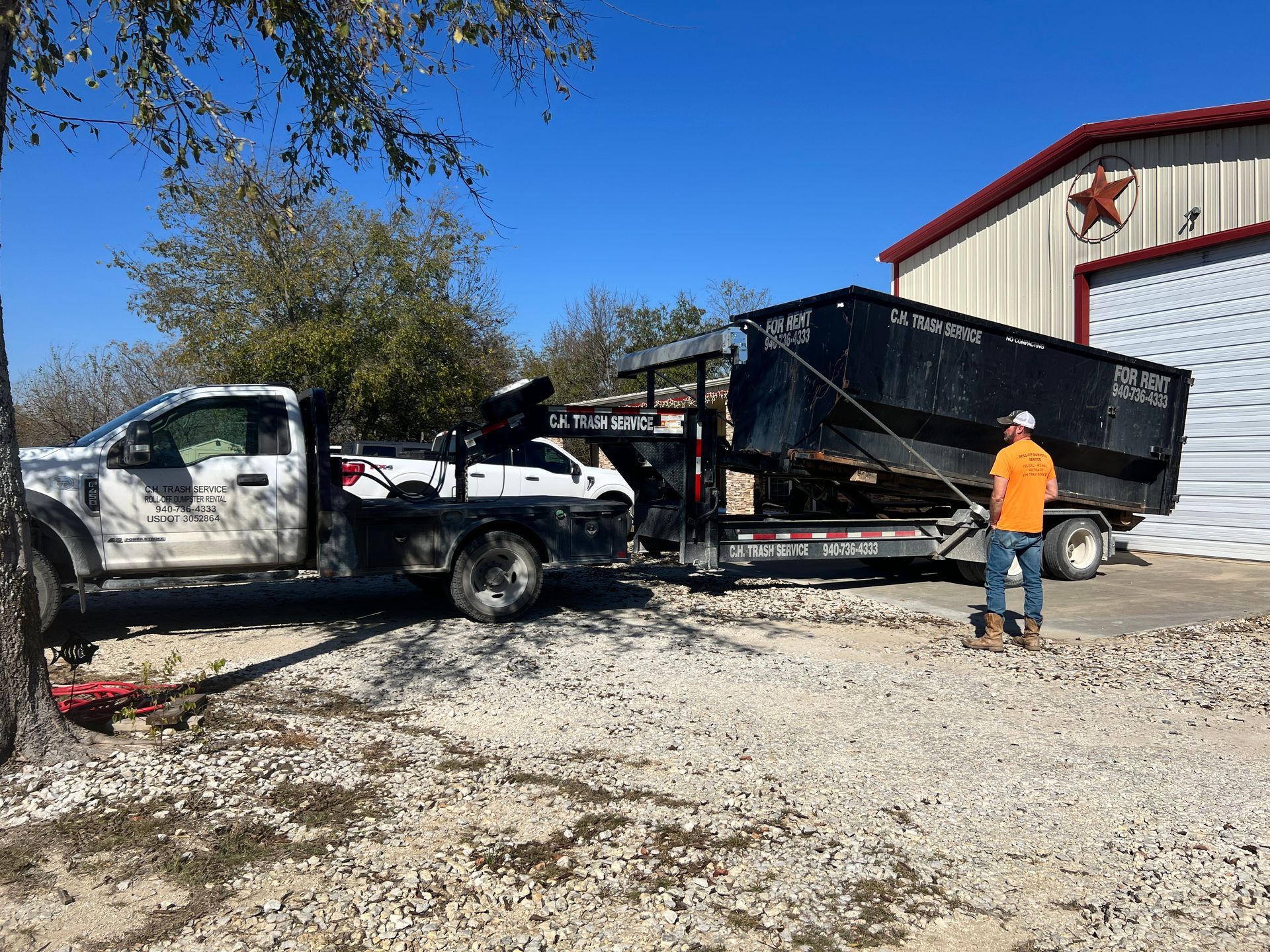White pickup truck towing a trailer with a large dumpster; a person standing near it. Sunny, outdoor setting.