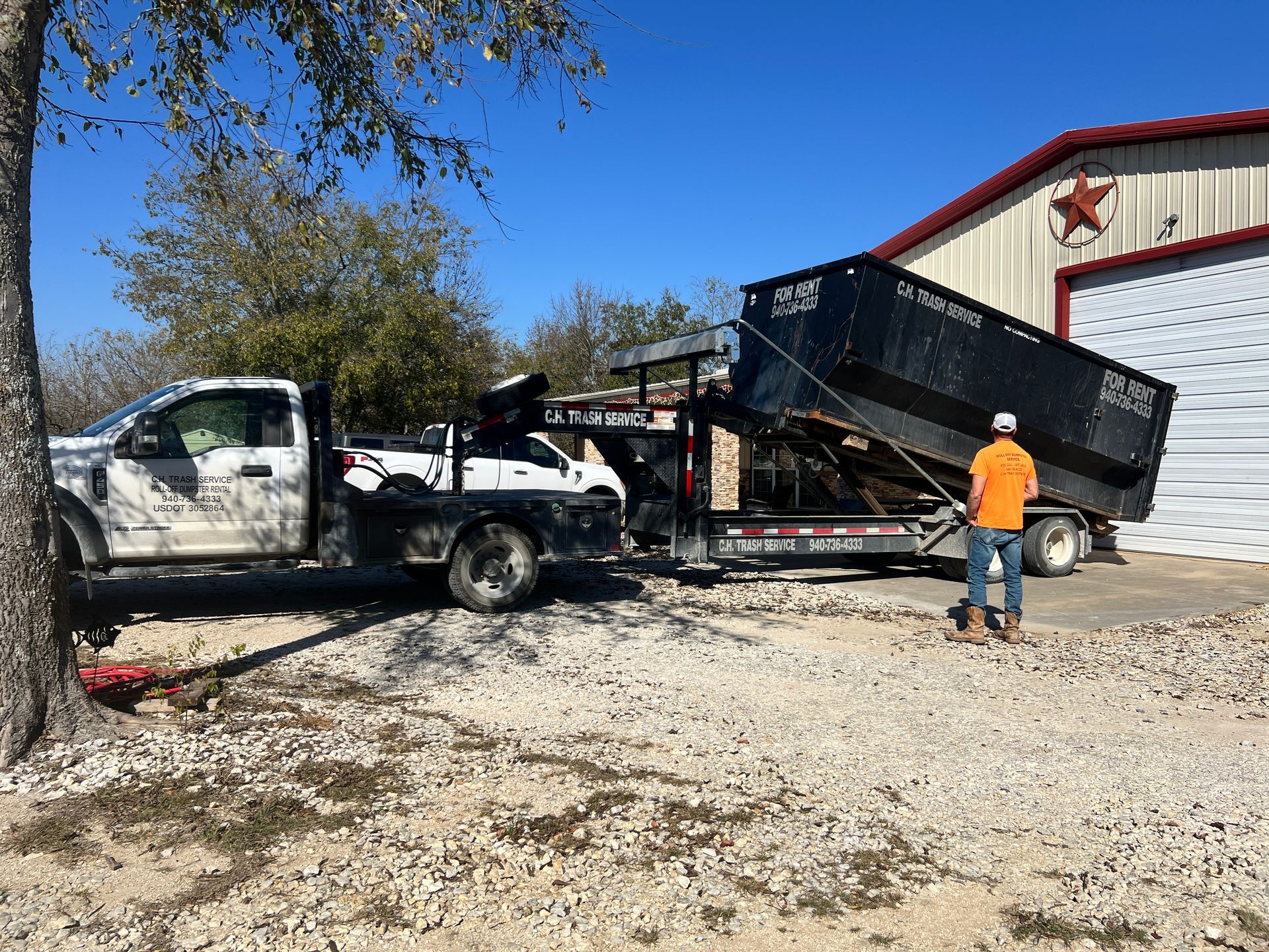 Truck hauling a trailer with a dumpster, man standing nearby, building in the background.