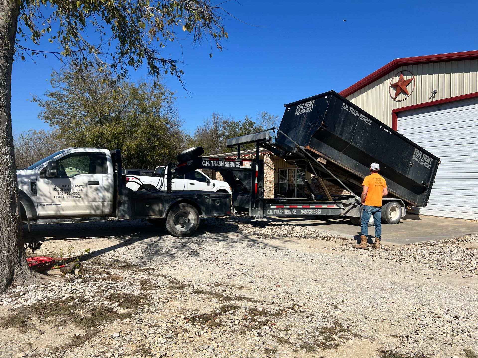 Truck with trailer dumping a large container at a building. A person in orange vest watches. Gravel driveway.