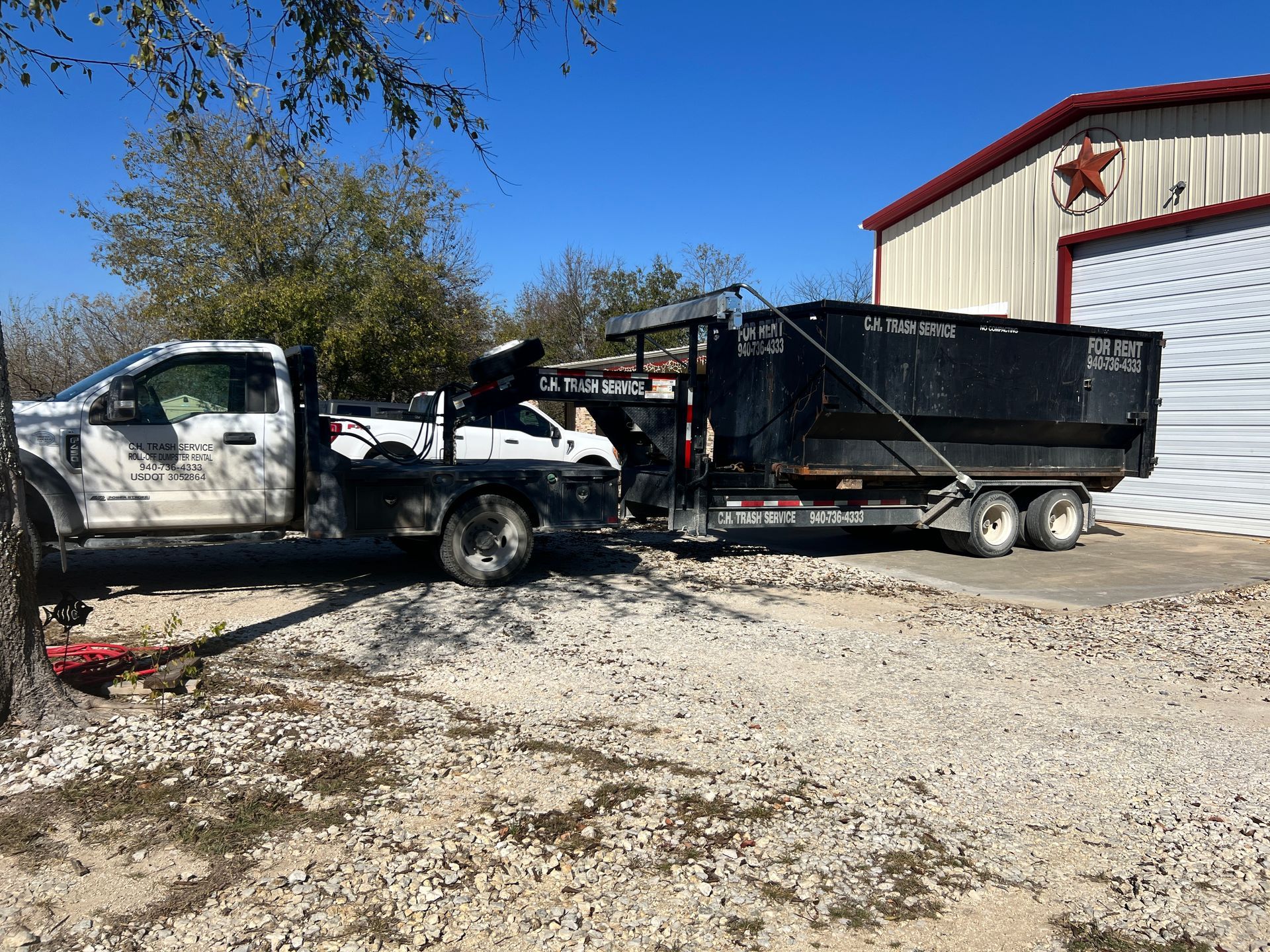 White truck towing a flatbed with a car and dumpster trailer in front of a white building with a red star.
