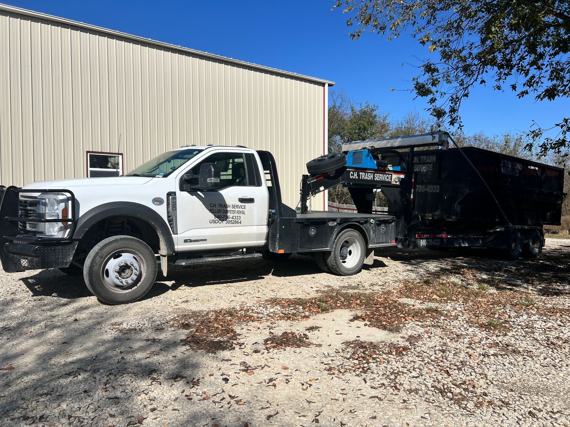 White flatbed truck towing a black trailer in front of a tan building under a blue sky.