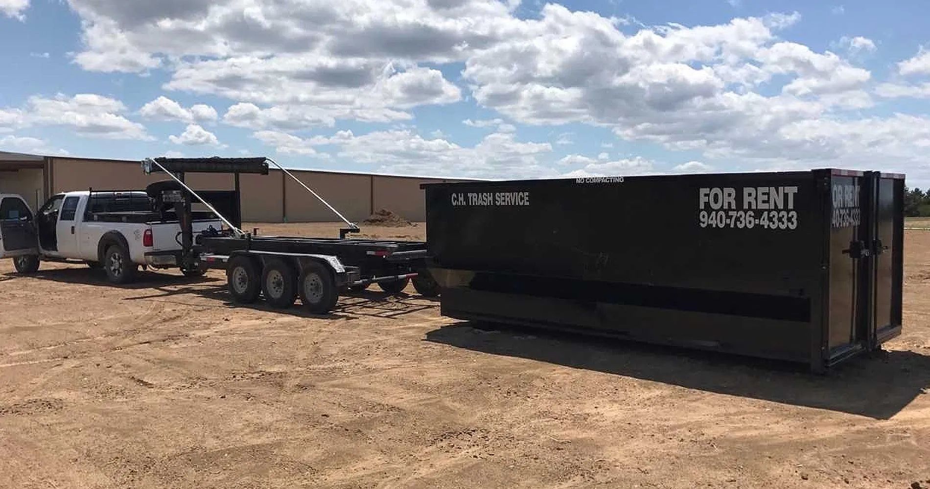 A black dumpster is being loaded onto a trailer with a truck in a dirt lot under a cloudy sky.