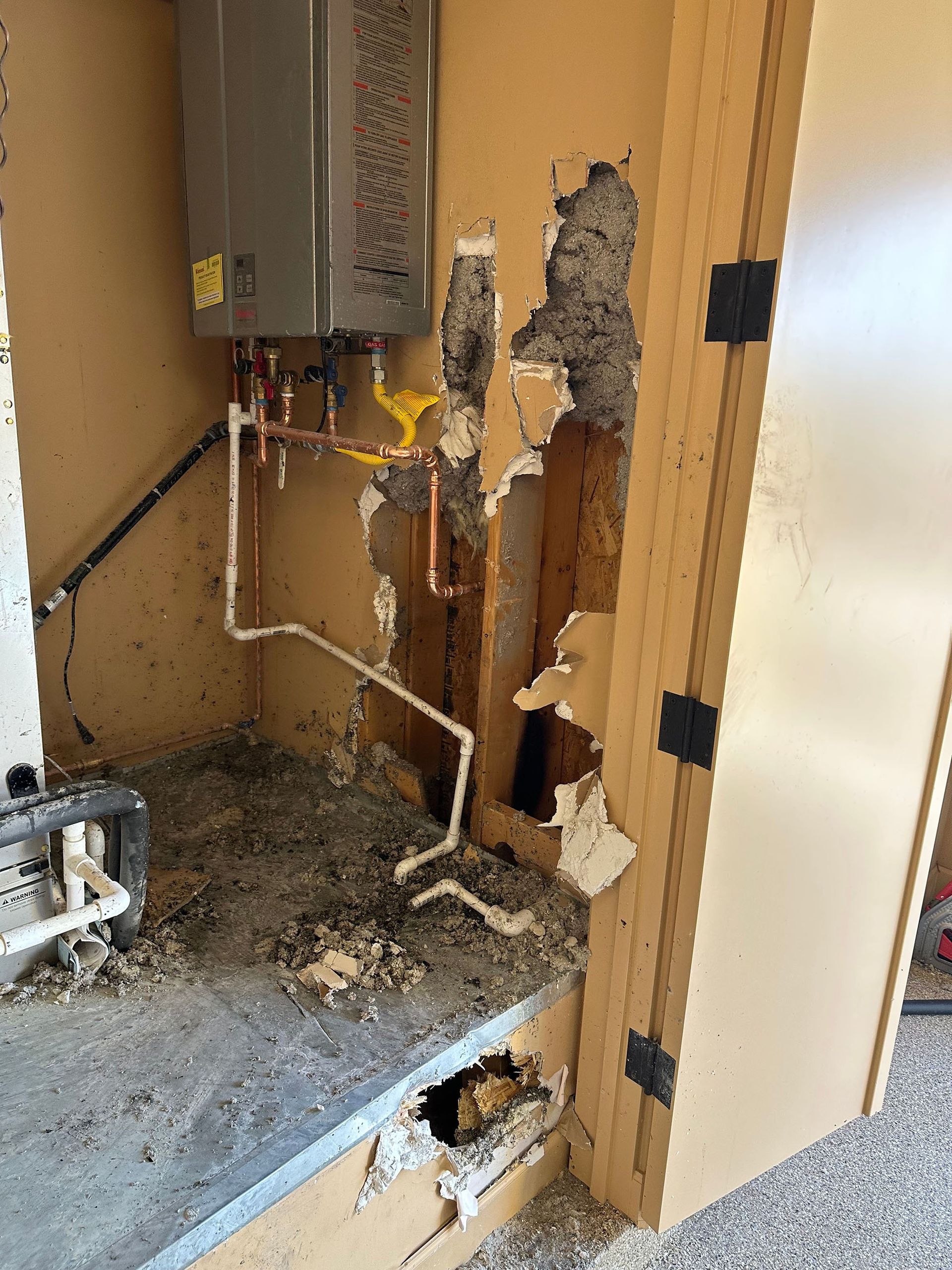 A water heater is sitting in a corner of a room with a hole in the wall.