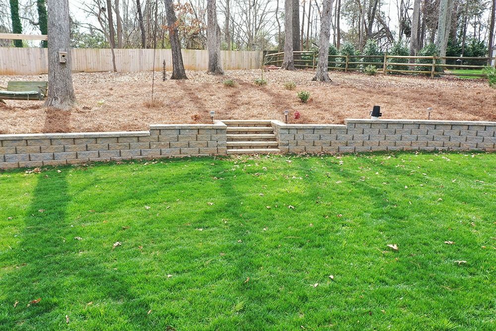 Green lawn with stone retaining wall, steps, and bare trees in a wooded backyard