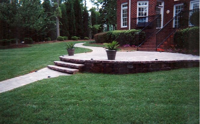 Curved stone patio and steps beside a brick house, with a potted plant and green lawn