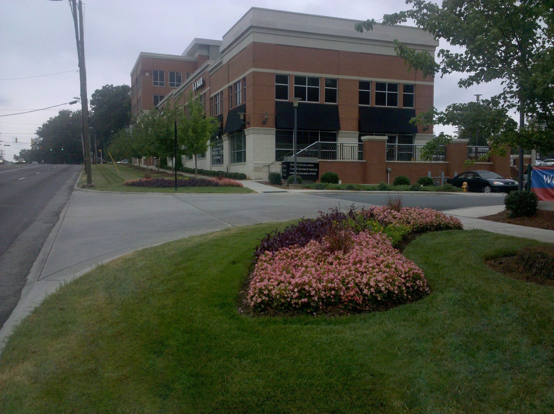 Street corner with brick office building, curved sidewalk, and pink flower bed under cloudy sky