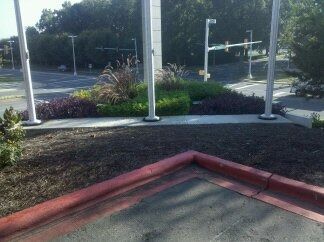 Outdoor sidewalk with red curb, white poles, and landscaped median near a road intersection