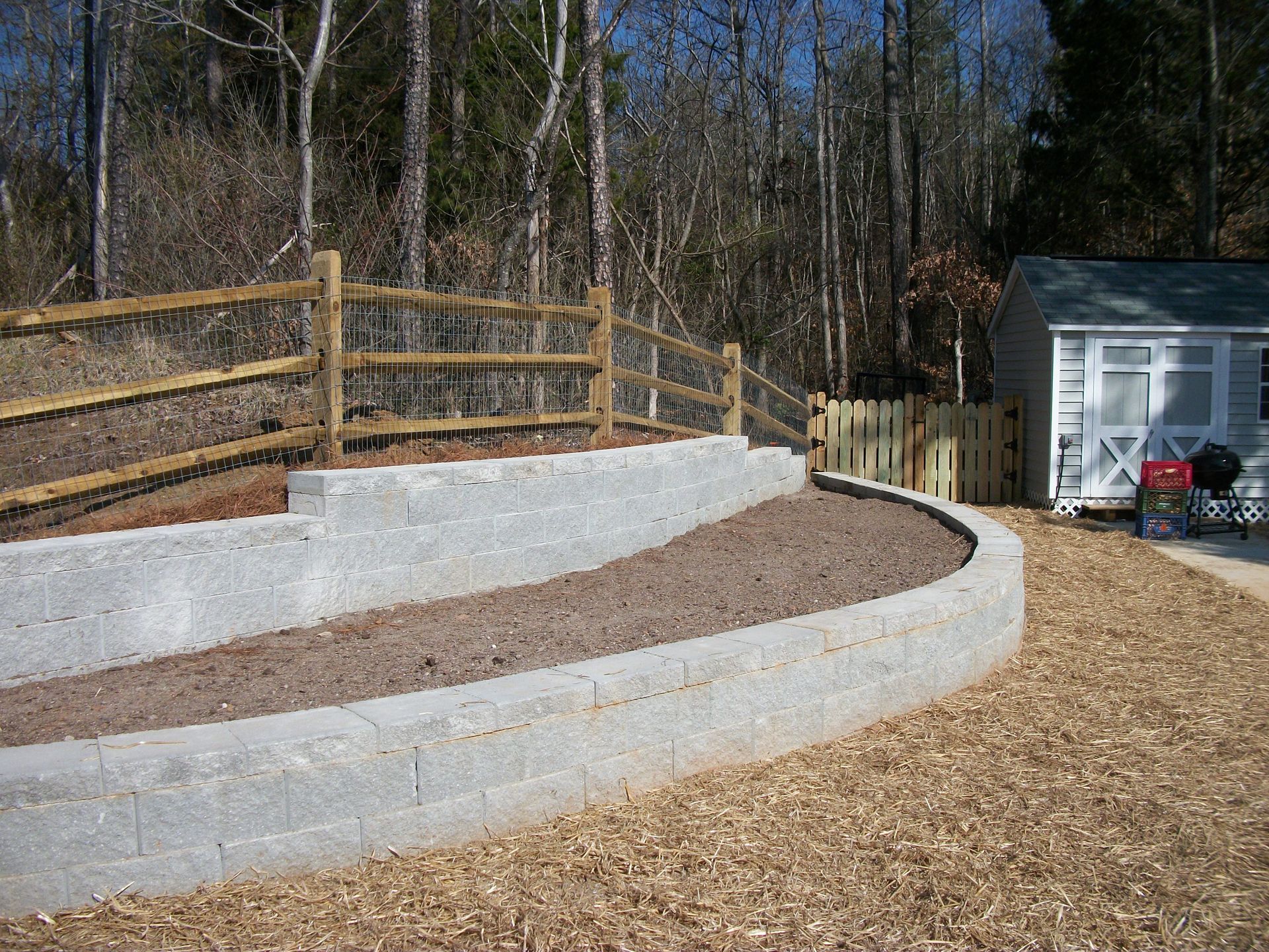 Curved white retaining wall beside a wood fence and gravel yard, with a small shed in the background.